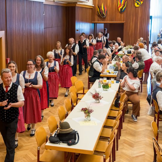 Eine Gruppe von Menschen ist in einem Saal und trägt traditionelle Kostüme, klatscht. Lange Tische sind aufgestellt, mit Blumen und Getränken. Der Raum hat Holzwände und -böden.