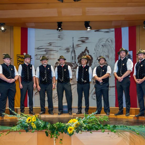 Sieben Männer in traditioneller Kleidung, einschließlich Hüten und Medaillen, stehen auf einer Bühne mit Blumen und blicken in die Kamera. Dahinter zeigt eine Kulisse eine Kirche mit einer Flagge.