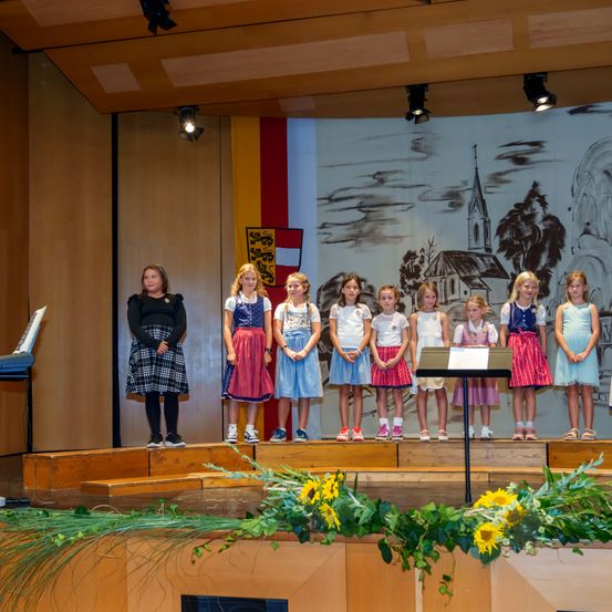 Eine Gruppe von Kindern in traditioneller Kleidung steht auf der Bühne und schaut ins Publikum. Eine Frau steht links und hält eine Musiknote. Im Hintergrund befindet sich eine Kirche, Bäume und eine Flagge.