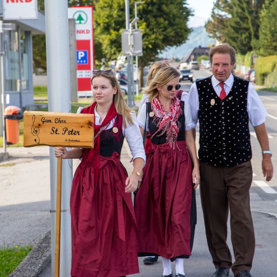 Drei Menschen in traditioneller bayerischer Kleidung gehen eine Straße entlang, wobei im Vordergrund ein Schild mit der Aufschrift 'Gem. Char St. Peter' steht.