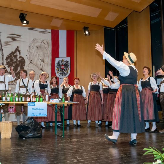 Eine Gruppe von Menschen in traditioneller Kleidung, die auf einer Bühne performt, mit Bierflaschen auf einem Tisch vor ihnen. Dahinter ist eine Wand mit einem Gemälde und einer Flagge.