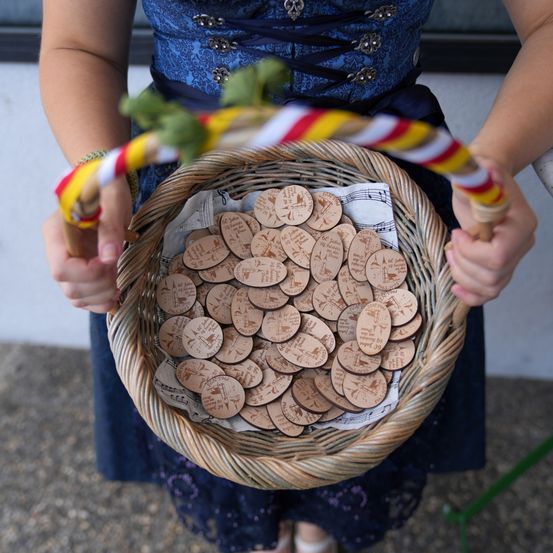 Eine Person in einem blauen Kleid hält einen Korb mit einem Griff, der zahlreiche hölzerne ovale Objekte mit Gravuren enthält. Der Korb liegt auf dem Schoß der Person.