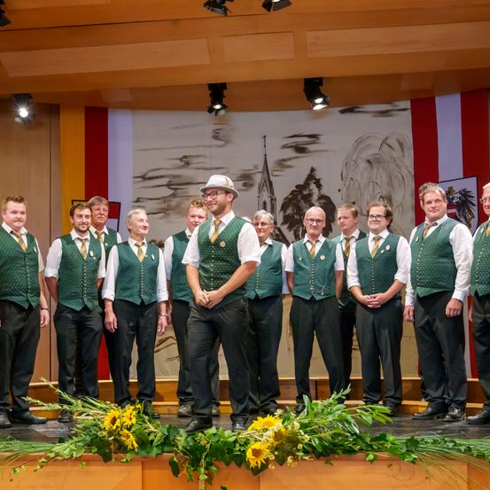 Eine Gruppe von Männern in grünen Westen und schwarzen Hosen steht auf einer Bühne mit Sonnenblumen davor. Sie alle lächeln und schauen in die Kamera. Hinter ihnen ist eine Kulisse mit einem Dorfgemälde.