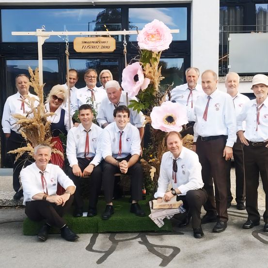 Eine Gruppe von Männern und Frauen in weißen Hemden und roten Krawatten posiert für ein Foto vor einem Gebäude mit einem Schild, das 'Singeheinschaft Feistritz/Drau' besagt.