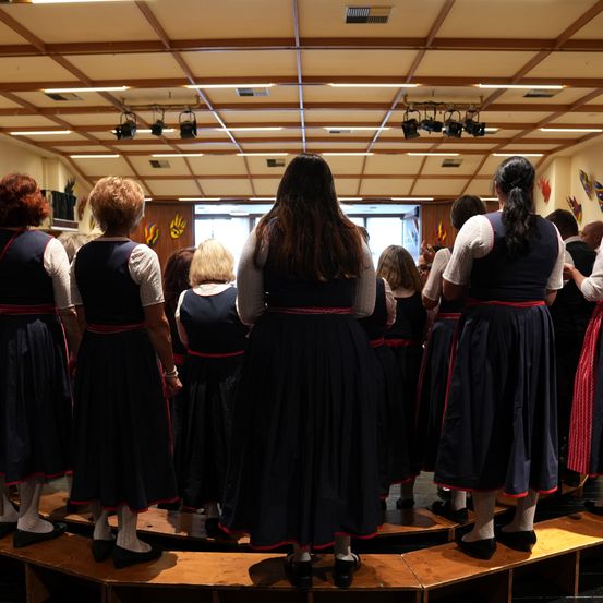 Eine Gruppe von Frauen in traditionellen Kleidern steht auf einer Bühne mit Holzboden. Der Raum hat Holzbalken an der Decke, und die Wände sind mit farbenfrohen Gemälden verziert. Die Frauen schauen dem Publikum zu, und der Raum ist gut beleuchtet.