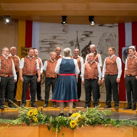Ein Männerchor steht auf einer Bühne mit einer Frau davor, die ein blaues Kleid trägt und ihnen gegenübersteht. Sie tragen passende Outfits mit roten und weißen Mustern. Ein bemalter Hintergrund ist hinter ihnen.