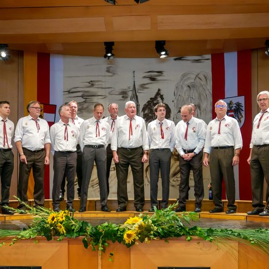 Eine Gruppe von Männern in gleichen Outfits steht auf einer Bühne, wahrscheinlich singend. Sie tragen weiße Hemden, rote Krawatten und dunkle Hosen. Hinter ihnen ziert ein Wandbild den Hintergrund.