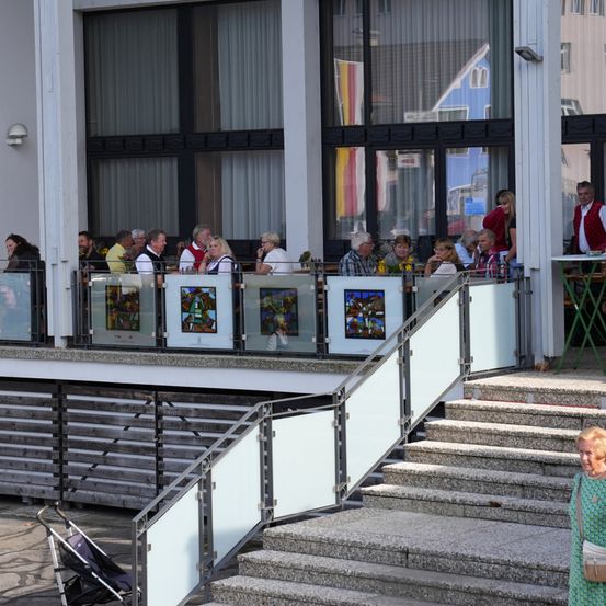 Eine Gruppe von Menschen sitzt an Tischen auf einer Außenterrasse eines Gebäudes mit Glaswänden, Geländern und einer Flagge. Eine Frau geht die Treppe hinauf.