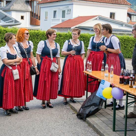 Fünf Frauen in traditioneller deutscher Kleidung stehen neben einem Tisch mit Getränken. Sie lächeln und unterhalten sich. Hinter ihnen steht eine Hecke und Häuser. Eine Frau hält eine Flasche.