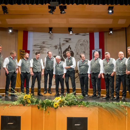 Ein Chor in traditioneller deutscher Kleidung steht auf der Bühne und schaut das Publikum an. Die Bühne ist mit Sonnenblumen und Grün geschmückt. Dahinter zeigt eine Wandmalerei eine Kirche und Berge. Scheinwerfer sind an der Decke montiert.