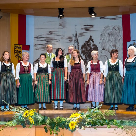 Eine Gruppe von Menschen in traditioneller Kleidung steht auf der Bühne. Sie werden von zwei Männern in formeller Kleidung begleitet. Die Bühne ist mit einer Kulisse eines Dorfes und einer Flagge geschmückt.