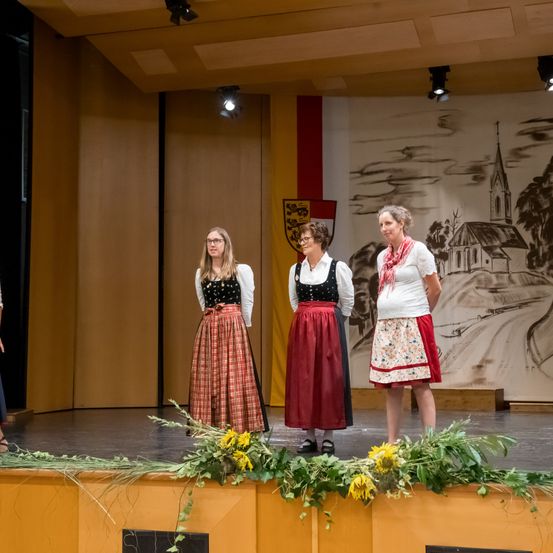 Drei Frauen in traditioneller Kleidung stehen auf einer Bühne, möglicherweise für eine Aufführung. Die Bühne ist mit Blumen und grünen Pflanzen dekoriert. Dahinter befindet sich eine Kulisse mit einem Gemälde einer Kirche und eines Dorfes.