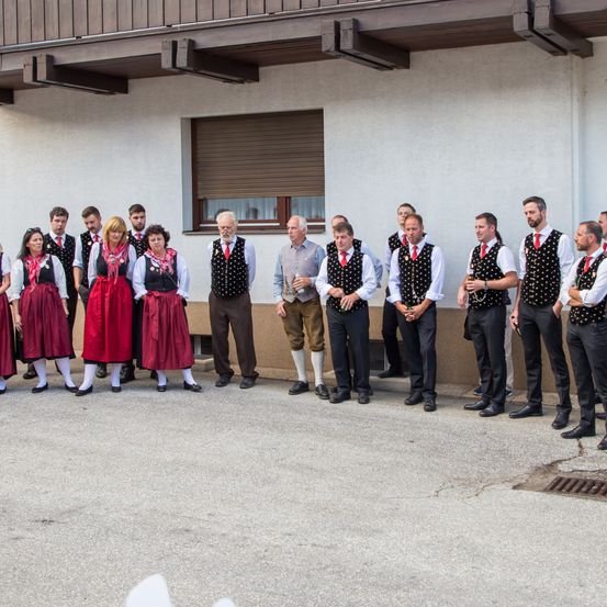 Eine Gruppe von Menschen in traditioneller Kleidung steht vor einem Gebäude. Die Gruppe besteht aus Männern und Frauen, die alle rote und weiße Schals tragen.