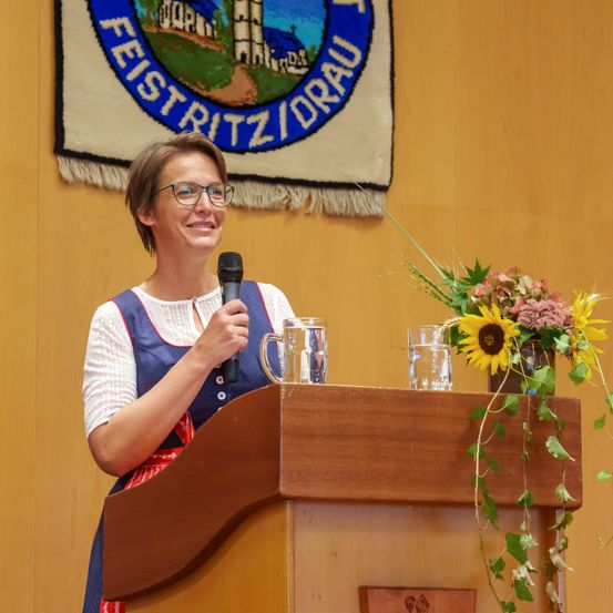 Eine Frau mit Brille spricht in ein Mikrofon an einem Podium mit Blumen an der Wand hinter ihr.