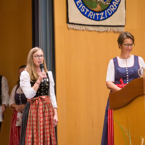 Eine Frau spricht in ein Mikrofon, während eine andere Frau vor einem Podium an einer gelben Wand mit einem Banner steht. Dahinter stehen zwei weitere Frauen in traditioneller Kleidung.