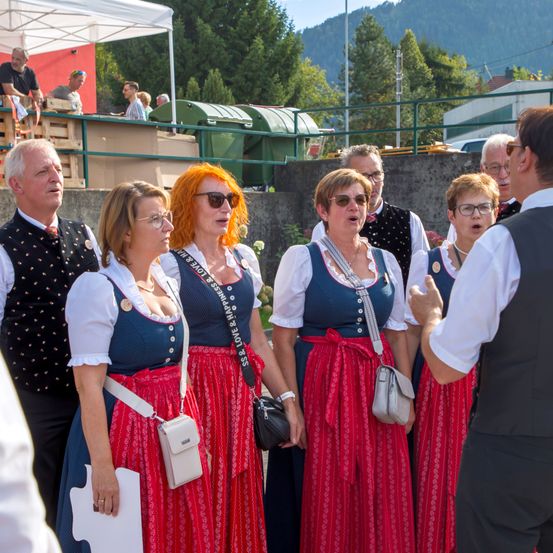 Eine Gruppe von Frauen in traditionellen Kostümen steht in einer Reihe, möglicherweise singend, während ein Mann zu ihnen spricht. Dahinter befindet sich ein Zaun mit einem Zelt, und mehrere Personen beobachten von der anderen Seite.