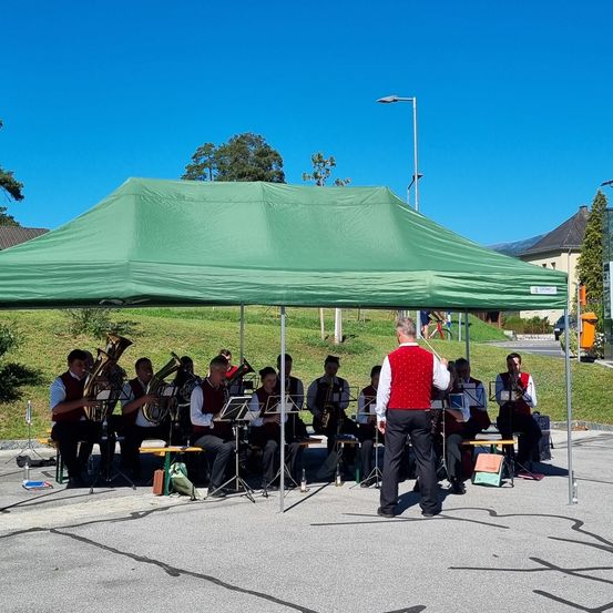 Eine Band tritt an einem sonnigen Tag unter einem grünen Zelt auf einer gepflasterten Oberfläche auf. Sie ist von Bäumen und Gebäuden im Hintergrund umgeben.