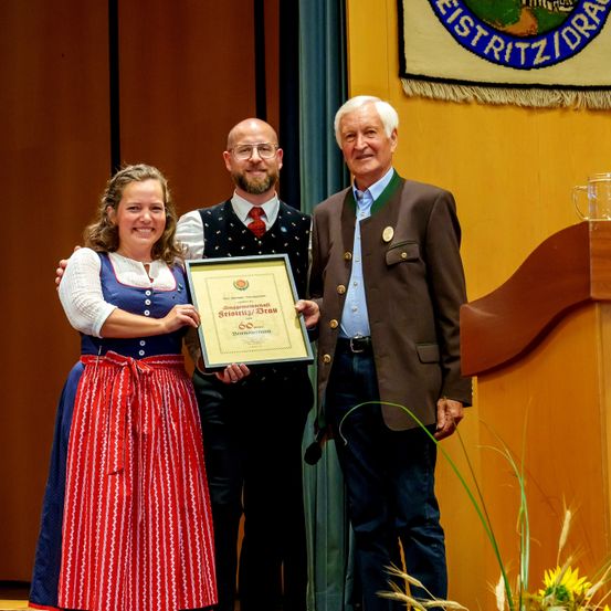 Eine Frau und zwei Männer halten ein gerahmtes Zertifikat vor einem Podium und lächeln. Der Mann auf der rechten Seite trägt einen braunen Anzug und der Mann auf der linken Seite trägt ein Weste und eine rote Krawatte. Eine blaue Flagge hängt an der Wand hinter ihnen.