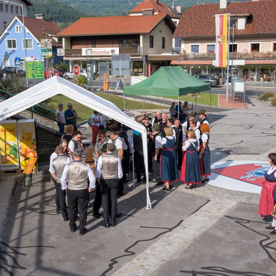 Eine Gruppe von Menschen in traditioneller Kleidung steht unter einem Zelt in einem Außenbereich, mit Gebäuden und Bergen im Hintergrund.