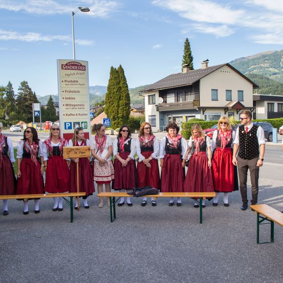 Eine Gruppe von Frauen in traditioneller Kleidung steht vor einem Restaurant mit einem Schild, das 'Sandriesser' steht. Sie posieren für ein Foto mit Bergen im Hintergrund.