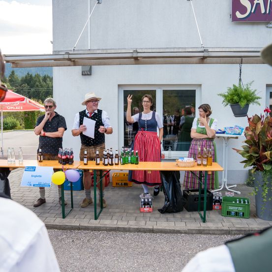 Eine Gruppe von Menschen steht hinter einem Tisch mit Flaschen, trägt traditionelle deutsche Kleidung, mit Pflanzen und Luftballons im Vordergrund.