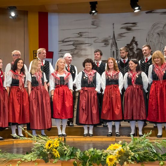Eine Gruppe von Menschen, sowohl Männer als auch Frauen, stehen auf der Bühne und tragen traditionelle rote Kleider und Schals, mit einem Hintergrund von einem Gemälde.