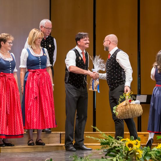 Eine Gruppe von Menschen in traditionellen Kostümen steht auf der Bühne. Zwei Männer schütteln sich die Hände. Einer der Männer hält einen Korb mit Brot. Die Frau auf der linken Seite trägt einen roten Rock.