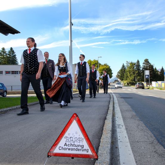 Eine Gruppe von Menschen in traditioneller Kleidung geht am Straßenrand, mit einem Warndreieck, das auf eine Chorwanderung hinweist.