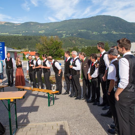 Eine Gruppe von Männern in traditioneller Kleidung steht im Freien, in der Nähe von Tischen und Bänken, mit Bergen im Hintergrund.