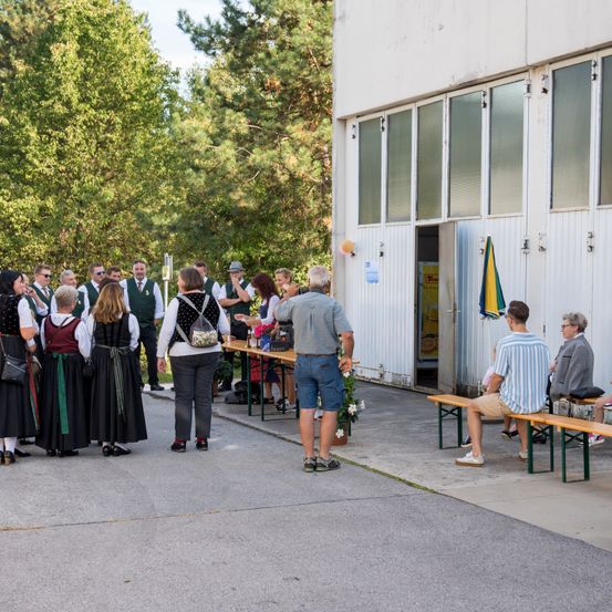 Eine Gruppe von Menschen in traditioneller Kleidung steht vor einem Gebäude mit offener Tür und einem Regenschirm. Eine Frau mit Rucksack und ein Mann in einem grauen Shirt sind dabei.