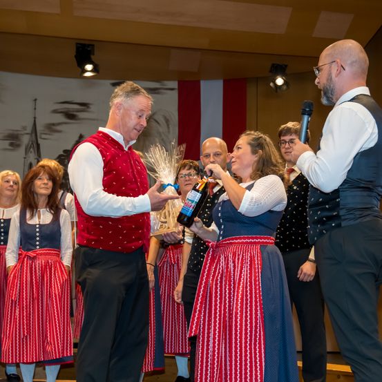 Eine Preisverleihung mit einer Gruppe von Menschen in traditioneller Kleidung, ein Mann hält eine Flasche und eine Frau empfängt sie. Ein Mann mit einem Mikrofon steht in der Nähe.