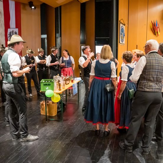 Eine Gruppe von Erwachsenen, gekleidet in traditionelle bayerische Kleidung, versammelt sich um einen Tisch mit Bierflaschen und Luftballons. Sie unterhalten sich und einige lächeln.