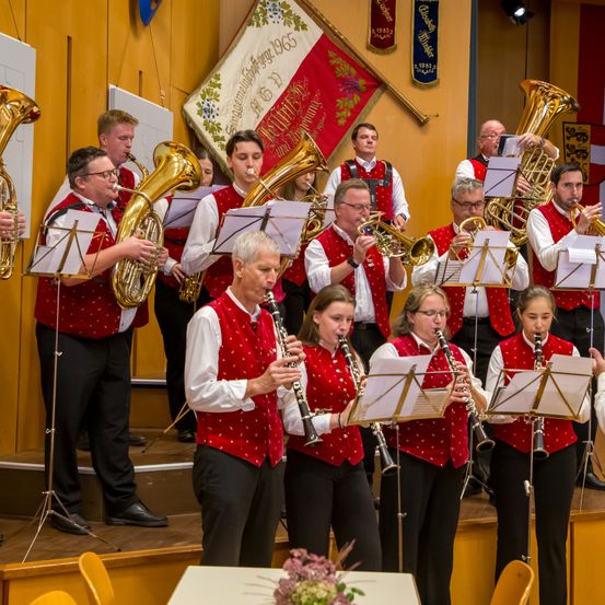 Eine Gruppe von Musikern in roten Westen spielt auf der Bühne ihre Instrumente. Einige spielen Blechblasinstrumente, andere Klarinetten. Hinter ihnen hängen zwei Fahnen an der Wand.