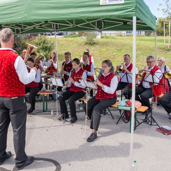 Eine Band spielt im Freien unter einem grünen Zelt. Die Mitglieder sitzen auf Bänken und spielen verschiedene Instrumente. Ein Mann steht vorne und leitet die Aufführung.