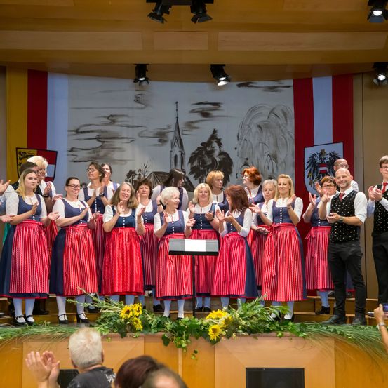 Ein Chor in traditioneller Kleidung tritt auf einer Bühne mit Blumen im Vordergrund auf. Ein Banner mit einer Kirchenmalerei im Hintergrund. Das Publikum klatscht.
