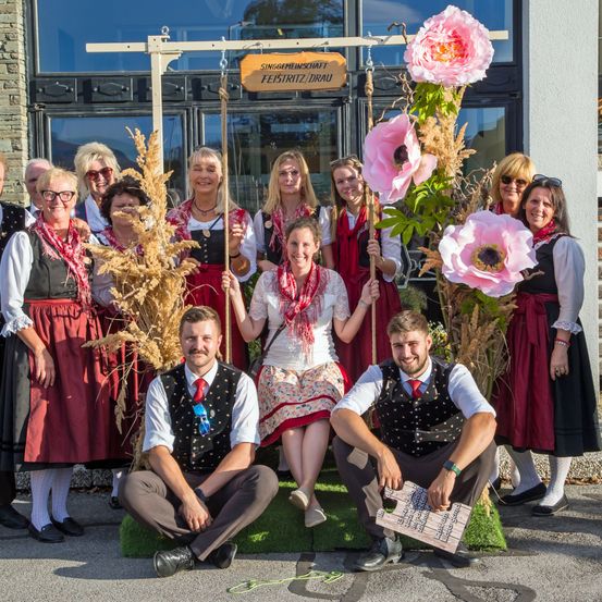 Eine Gruppe von Menschen in traditioneller deutscher Kleidung posiert für ein Foto vor einem Gebäude. Einige sitzen auf einer Schaukel mit großen rosa Blumen, während andere hinter ihnen stehen. Ein Schild lautet 'Singgemeinschaft'.