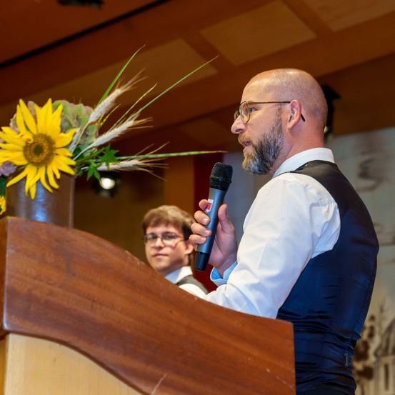 Ein Mann am Rednerpult mit Mikrofon, trägt eine Weste und Brille, mit einer Sonnenblume im Hintergrund.