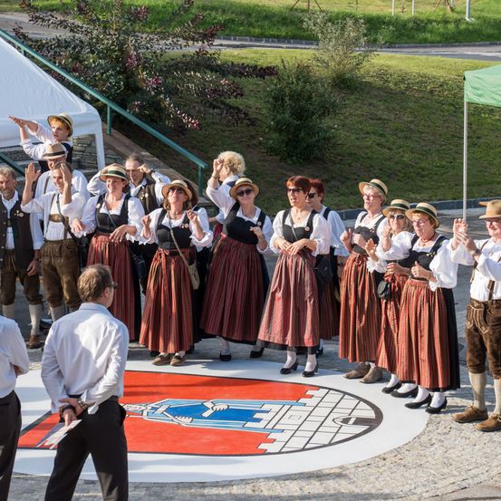 Eine Gruppe Erwachsener in traditioneller Kleidung klatscht und steht auf einem weißen kreisförmigen Muster mit einem Logo. Ein Mann in einem weißen Hemd hält ein Papier.