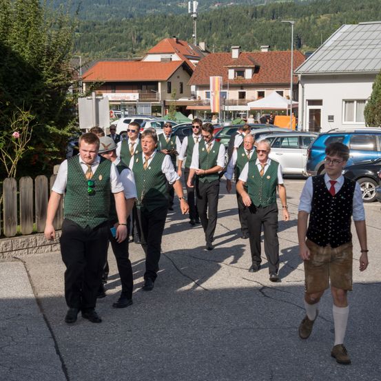 Männer in traditioneller Kleidung gehen eine Straße entlang mit geparkten Autos und Häusern im Hintergrund.