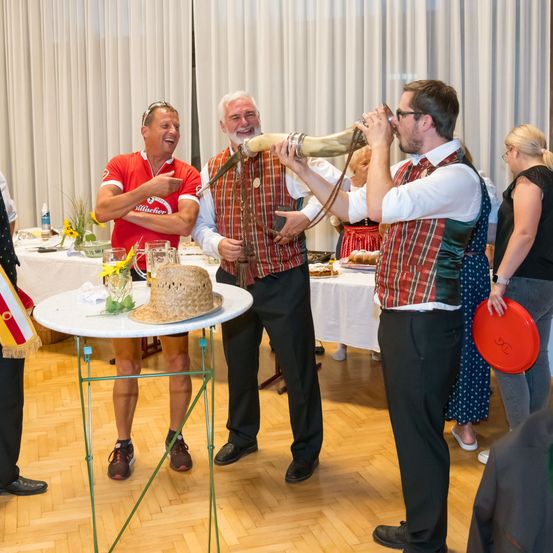 Ein Mann bläst ein Horn, während ein anderer Mann einen Hut auf einem Tisch hält, mit mehreren Leuten, die herumstehen, und Tischen mit Essen und Getränken im Hintergrund.