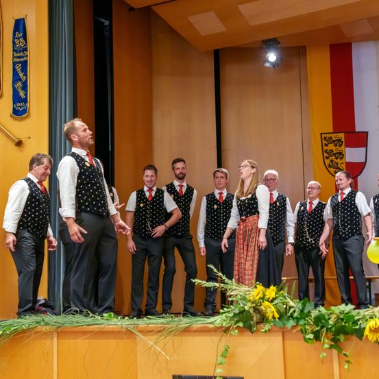 Eine Gruppe von Menschen, gekleidet in traditioneller Kleidung, steht auf einer Bühne mit einer Sonnenblume davor. Der Mann in der Mitte spricht, während andere zuhören. Hinter ihnen hängt eine Flagge und eine Wand mit Bannern.