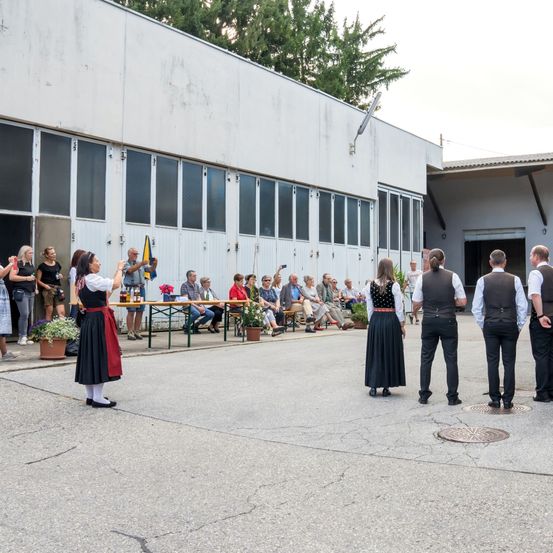 Eine Gruppe von Menschen ist im Freien versammelt, möglicherweise für eine traditionelle Veranstaltung. Eine Frau in traditioneller Kleidung steht vor einem Tisch, während andere auf Bänken und Tischen sitzen. Die Szene spielt sich vor einem Gebäude ab.