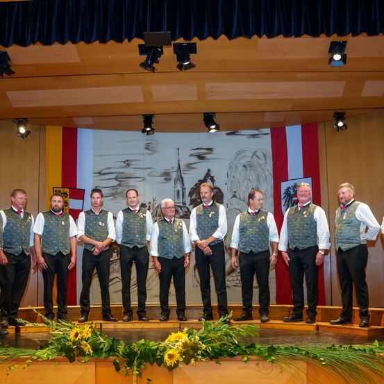 Neun Männer in identischen grünen Westen und weißen Hemden stehen auf einer Bühne vor einer Kulisse mit einer gemalten Szene einer Kirche und Bergen.