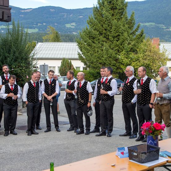 Eine Gruppe von Männern in traditioneller Kleidung steht im Freien. Sie halten Flaschen und Gläser. Im Hintergrund befinden sich Berge, Bäume und ein Gebäude.