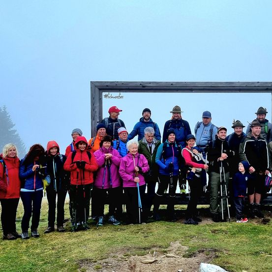 Eine Gruppe von Wanderern steht in einem Berggebiet, einige tragen Mützen und halten Trekkingstöcke. Hinter ihnen befindet sich ein Holzrahmen mit dem Hashtag #Schwarzee.