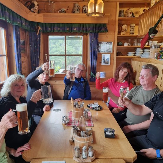 Eine Gruppe von Erwachsenen, alle lächelnd, sitzen um einen Holztisch in einer rustikalen Umgebung und heben ihre Getränke zum Anstoßen. Sie befinden sich in einem Raum mit Holzwänden und Regalen mit verschiedenen Gegenständen.