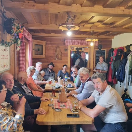 Eine Gruppe von Menschen sitzt in einer Holzhütte um einen Tisch herum. Einige unterhalten sich, während andere ihre Handys betrachten. Der Tisch ist mit Tassen, Gläsern und Tellern gedeckt. Eine Lampe hängt von der Decke, und Jacken hängen an der Wand.