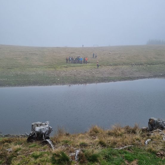 Eine Gruppe von Menschen steht in der Nähe eines Sees, mit einem nebligen Himmel und entfernten Bäumen. Einige tragen orangefarbene Westen, und eine einzelne Person steht am Wasserrand.