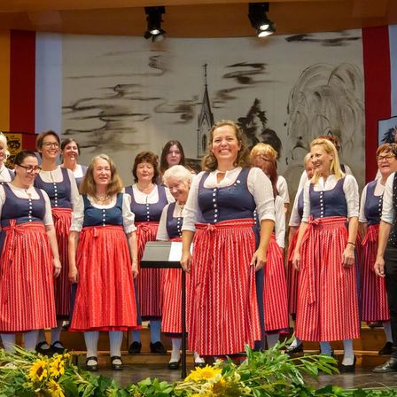 Eine Gruppe von Personen, einschließlich Frauen in traditioneller Kleidung und Männer in formeller Kleidung, steht auf der Bühne, möglicherweise singend. Vor ihnen sind Sonnenblumen und Grünpflanzen. Scheinwerfer sind an der Decke montiert.