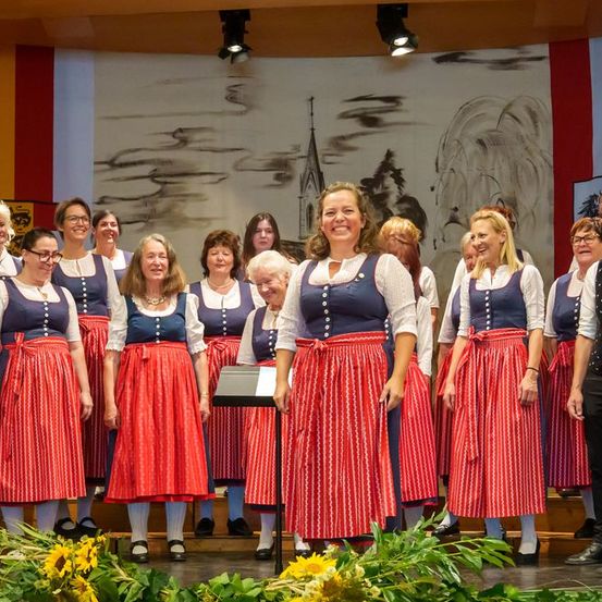 Eine Gruppe von Personen, einschließlich Frauen in traditioneller Kleidung und Männer in formeller Kleidung, steht auf der Bühne, möglicherweise singend. Vor ihnen sind Sonnenblumen und Grünpflanzen. Scheinwerfer sind an der Decke montiert.